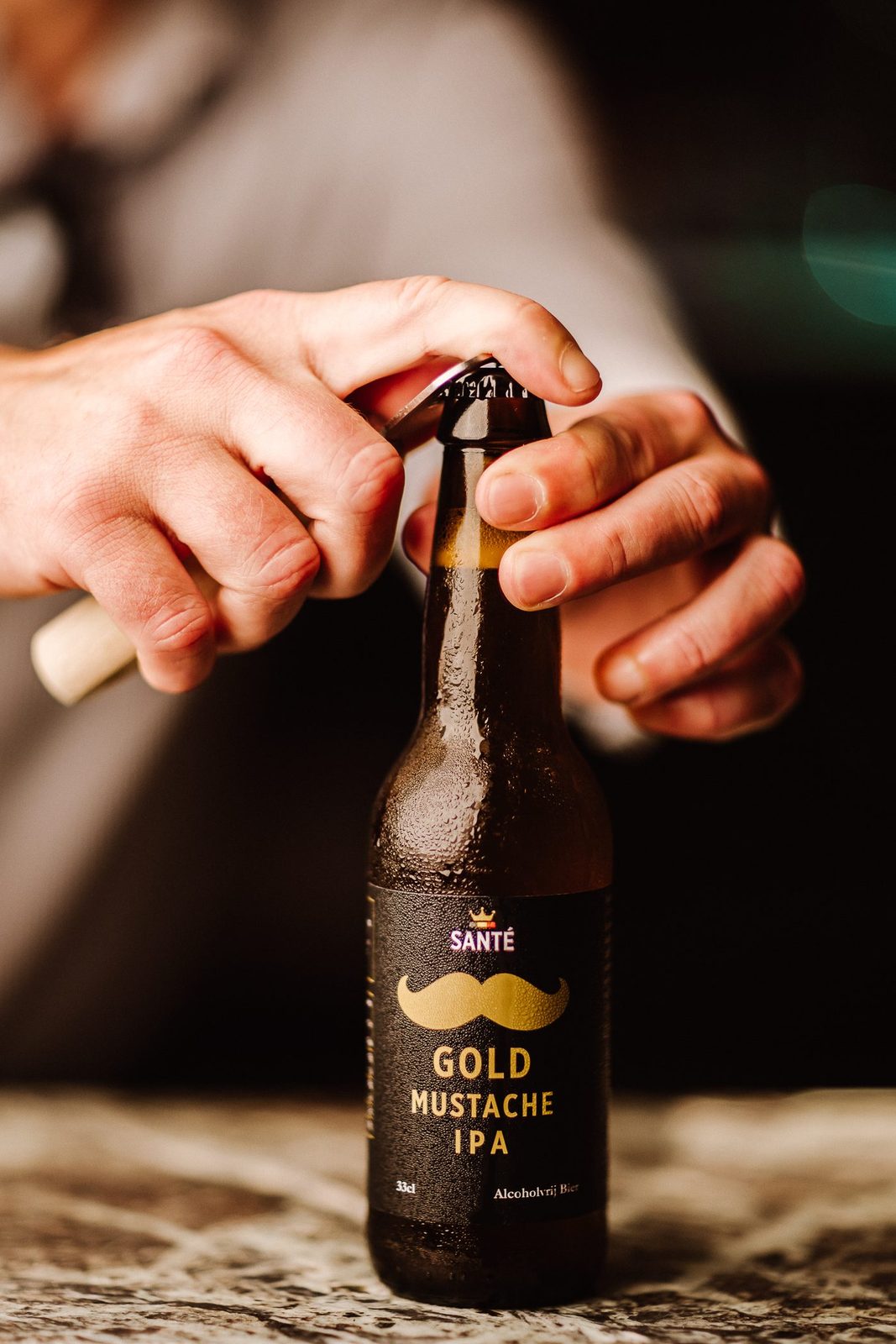 Une main ouvrant une bouteille de Gold Mustache IPA au décapsuleur, dehors au soleil