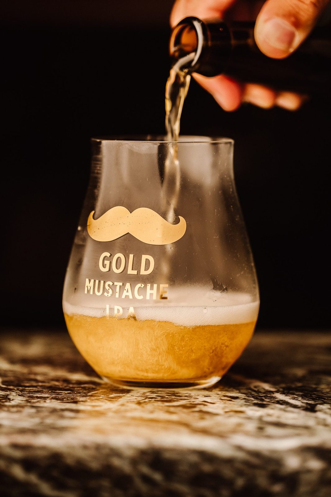 Une Gold Mustache IPA versée dans un verre tulipe siglé, sur une table en pierre