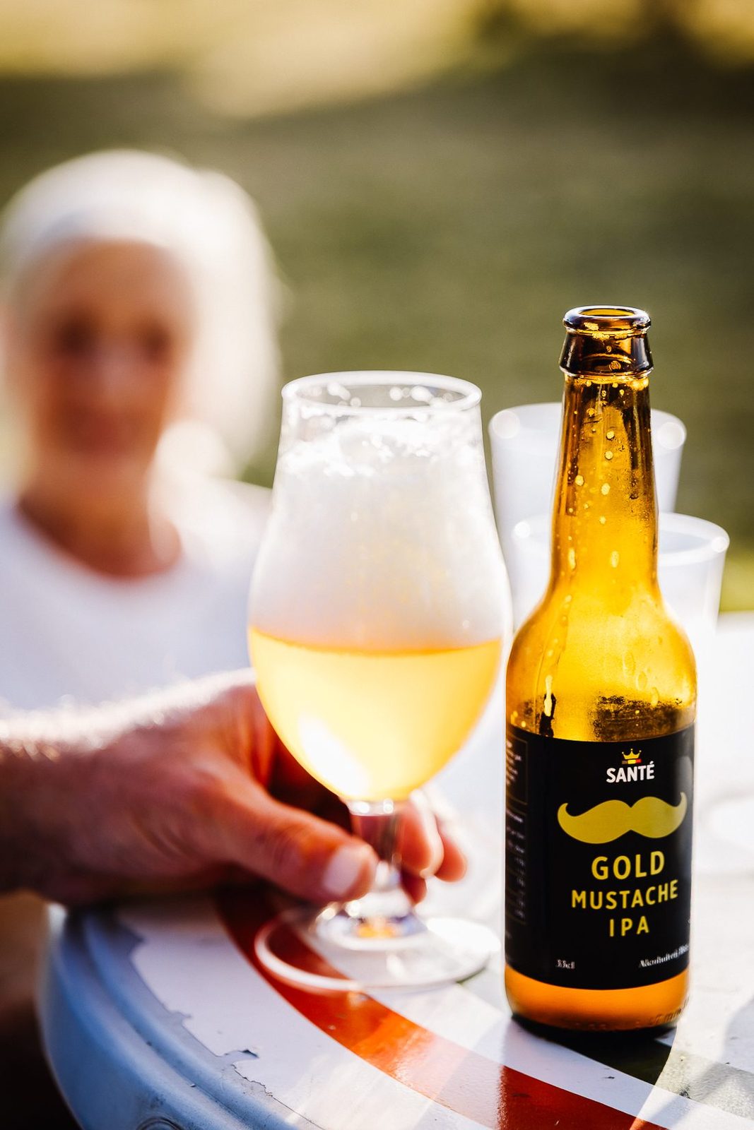A glass and a bottle of Gold Mustache IPA on a table outdoors, someone seated behind