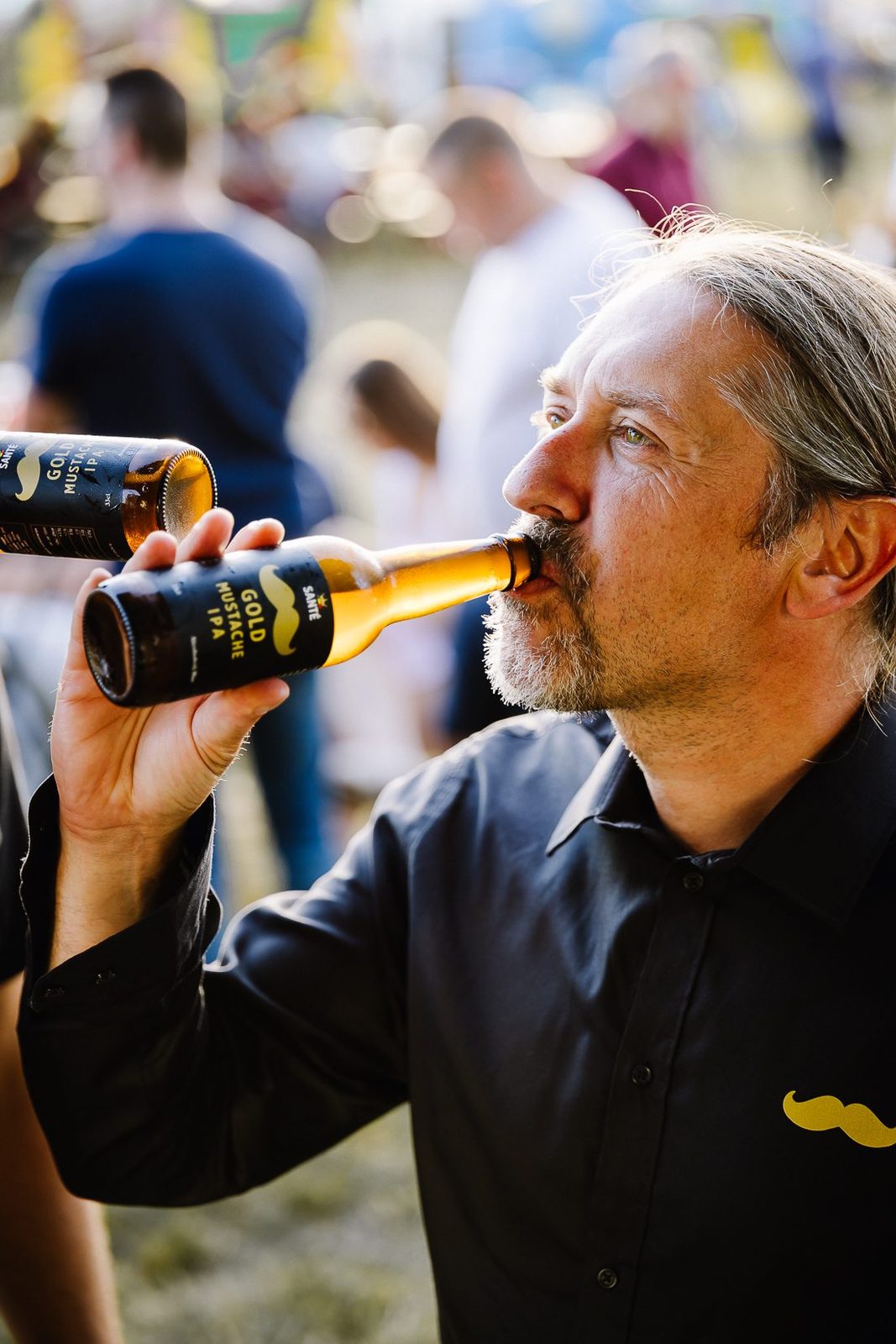A man drinking Gold Mustache straight from the bottle at a busy festival
