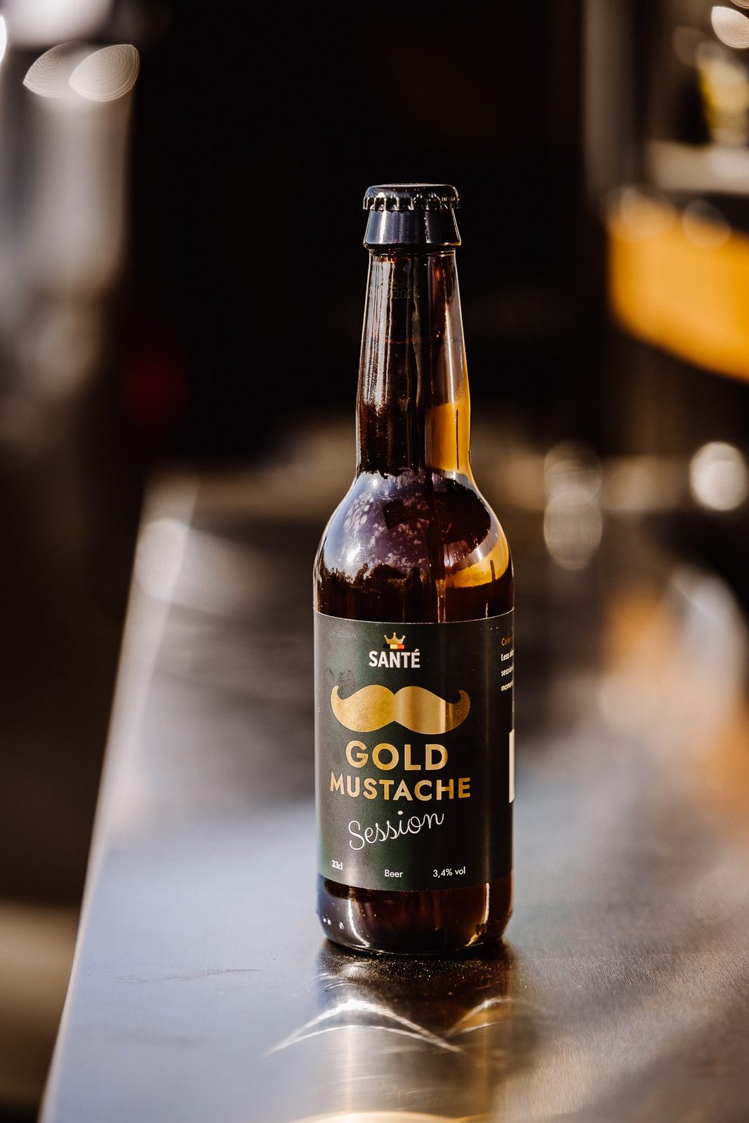 A bottle of Gold Mustache Session 3,4% standing on a stainless steel bar, warm light behind it